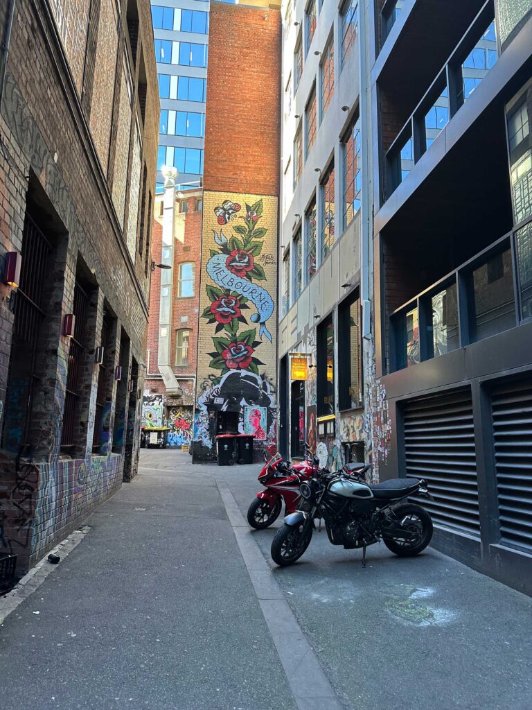 A narrow Melbourne laneway showcasing vibrant street art, including a large floral mural with 'MELBOURNE' text, surrounded by a mix of old brick and modern buildings, with two motorcycles parked in the foreground